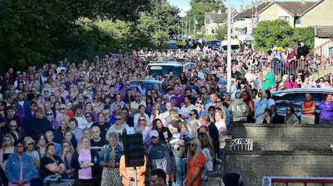 Pacemaker A large group of people, many wearing purple, stand in the street outside Sarah Montgomery's house. 