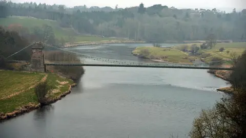 The river Tweed on a bend with the Union bridge, a narrow suspension bridge, in the foreground. On one side is a wooded forest, on the other fields