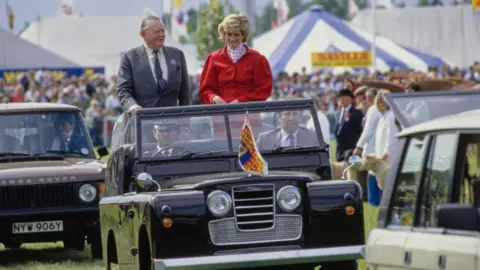 Getty Images A man and a woman stand in the back of an open top car as they move through a rural show. The man wears a grey suit while the woman is wearing a red jacket with a red and white polka dot blouse. 