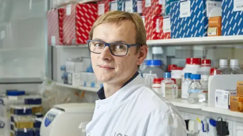 Graham is a professor of cancer research at a laboratory institute in front of the shelves full of tractor and boxes. He looks directly to the camera and wears a white laboratory coat and blond hair glasses