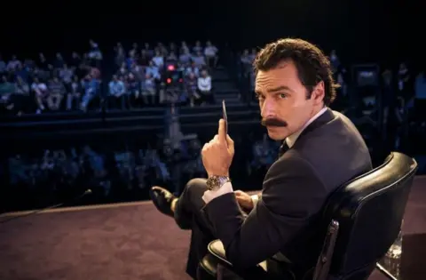 Actor Aidan Turner faces the camera in his role as a TV show host in the adaptation of Jilly Cooper's book Rivals. He is wearing a dark suit and has a thick moustache and is sitting in a stylish black leather chair with a TV audience visible, although not in focus, in the background