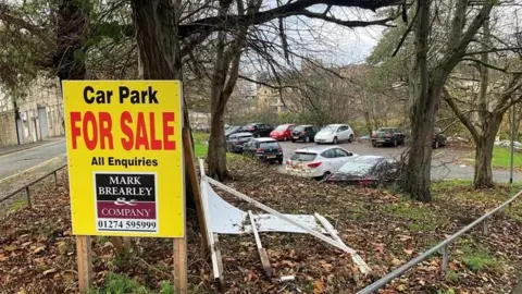 LDRS A yellow and black cap park for sale sign in a wooded area near a car park