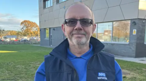 University Hospital Dorset David Carter smiling at the camera is wearing a royal blue jumper and navy blue NHS gilet. 