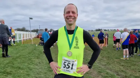 Wood wearing a yellow running vest, with a running track and other runners in the background, resting his hands on his hips, smiling into the camera. He is wearing a medal on a green strap around his neck.