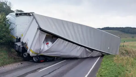 A large lorry lies on its side, completely blocking a road that sits between fields