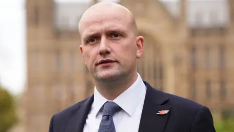 PA Media Stephen Flynn, a bald man wearing a dark blue suit and tie with a white shirt, stands in front of the Houses of Parliament 