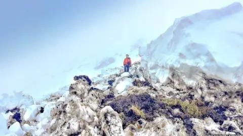 SAIS Southern Cairngorms An avalanche forecaster stands among avalanche debris. Some of the snow is mixed with grass, peat and soil the snow has dragged down during the slide.