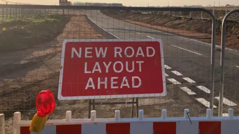 A road, with barriers in front, a large red sign and mounds of earth either side.