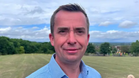 Calum Miller, a man with short dark hair, is pictured looking at the camera. He is wearing a blue checkered shirt with a collar and is stood outside during the day. Behind him there is a large field of grass, and beyond that a row of trees.