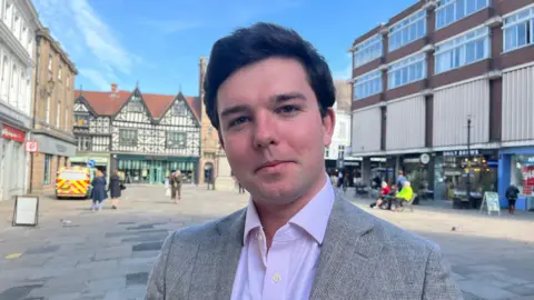 Alex looking into the camera; he is stood in Shrewsbury's town square, a large paved area with shoppers walking around in the background and shops around the edges. Alex is wearing a grey blazer and white collared shirt. The sky is bright blue and there is lots of sunshine. 