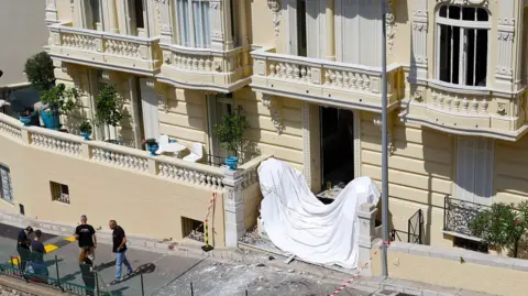 Frontage of a large cream coloured residential building in Monaco with damage to windows and a white sheet accross the front steps and police standing near the front of the building