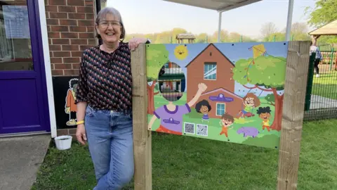 BBC/Lucy Ashton A woman with a grey bob is wearing glasses, a dark patterned blouse and jeans. She is stood smiling next to a sign with a brightly coloured cartoon of children playing and a hole where children can put their head through