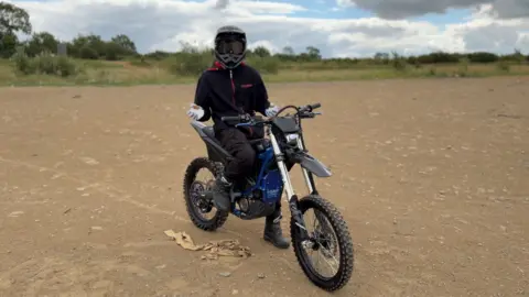 A person on a motorcycle wearing a helmet and dark clothing. They are in the middle of a piece of bare land, with a dusty gravel on the floor, and some low shrubs and trees in the background.