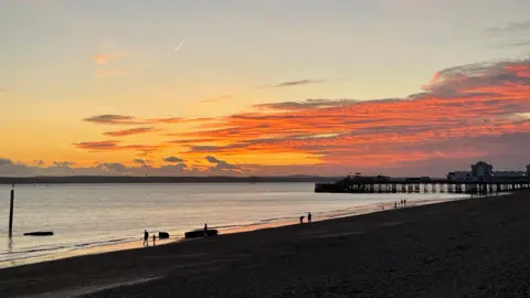 A golden sunset casts an orange and yellow glow across the sky over a beach with a pier sticking out into the water. Several people are walking along the beach.