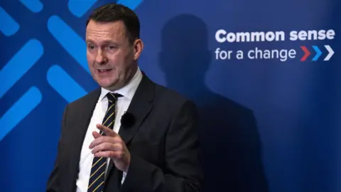 PA Media Russell Findlay, a man with dark hair, photographed in front a blue Conservative-branded sign. He is wearing a dark suit, a black and yellow striped tie and a white shirt. He is pointing with his left hand. 