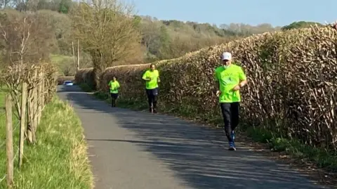 Javad Malik Three runners wearing high-vis yellow or green t-shirts, running on a narrow country lane, with a hill and trees in the background.