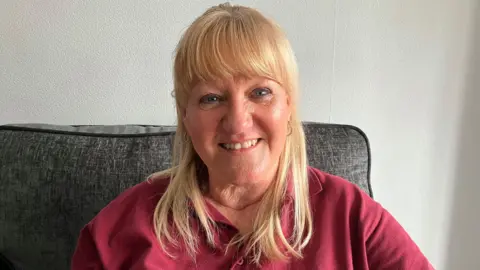 BBC/Jack Hadaway-Weller A woman with straight blonde hair is seated on a dark sofa against a textured white wall. She is wearing a red collared shirt and small hoop earrings, facing the camera directly.