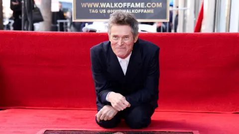 Reuters Willem Defoe sits on a red carpet as his unveiling on the Hollywood walk of fame takes place - he is is on his knees, leaning over a plaque on the walk