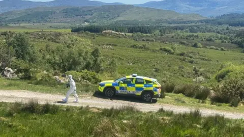 RTÉ A GARDA car parked at the side of a narrow dirt road. There is a person in a white forensic suit walking up the path away from the car. Rolling green hills are in the backdrop.