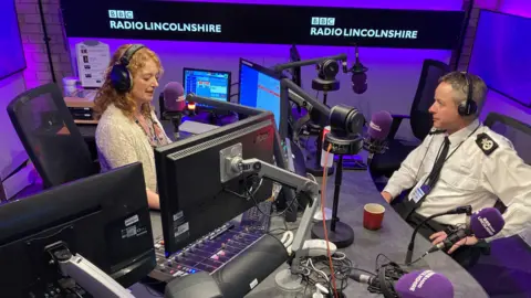 Frances and Paul are in a radio studio lit with purple lights. They sit at a grey desk, which has four computer screens, and five purple microphones. They are facing each other, and both are wearing black headphones. Frances has curly strawberry blonde hair and is wearing a cream cardigan. Paul has short dark hair with a tint of grey and is wearing a white police shirt and black tie.