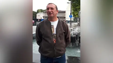 PSNI A picture of a man standing outside in front of a water feature. He has short hair and is wearing a white shirt under a brown jacket and jeans. His hands are behind his back.
