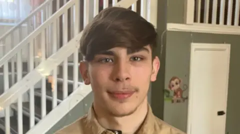 Grace Wood/BBC A young man with sweeping brown hair and very short facial hair smiles at the camera. He is wearing a beige shirt