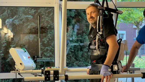 Anna Thomas Dan Richards using a machine to walk. He is standing and smiling, as he wears a harness and belts around his body.