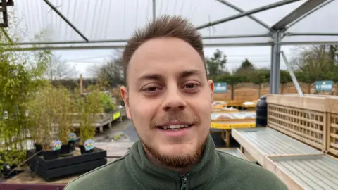 Jon Collins has short brown hair, a bit spikey at the top. He has a neatly trimmed brown beard and moustache and is wearing a green fleece. Behind him are flowers, plants and trees at a garden centre.
