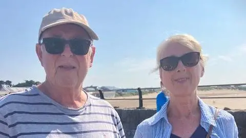 Peter and Anna Leivers are photographed from the shoulders up on Porthcawl seafront. The sandy beach can be seen behind them in the distance. Peter is wearing a grey striped t-shirt, black sunglasses and a beige cap. Anna is wearing a striped shirt, navy blue top and black sunglasses. She has blonde hair which is tied up.
