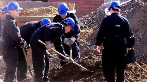 PA Media Police officers wearing black jumpsuits and blue helmets searching a building site, which has large piles of mud and rubble.