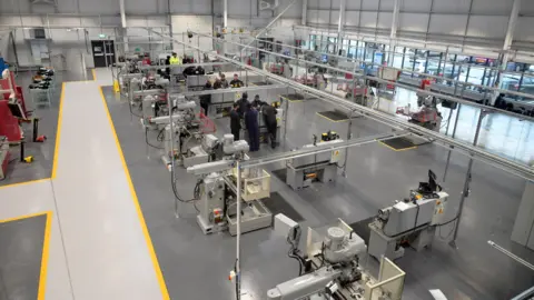 City of Wolverhampton College A huge factory with dozens of machines. It looks modern and smart. We can see cars parked outside through a window. There is a carved out safe area to walk with yellow lines either side. We can see some young people inside doing some work around the machines. They are wearing work clothing.