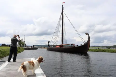 Peter Jolly/Northpix A man takes an image on his phone of replica Viking longship Draken Harald Hårfagre arriving at Inverness. There is a brown and white dog in the foreground.