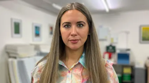 A close-up image of Sorcha Eastwood. She is a young woman, with long, highlighted hair. She is looking straight at the camera with a neutral expression on her face. She is wearing a rainbow coloured shirt.