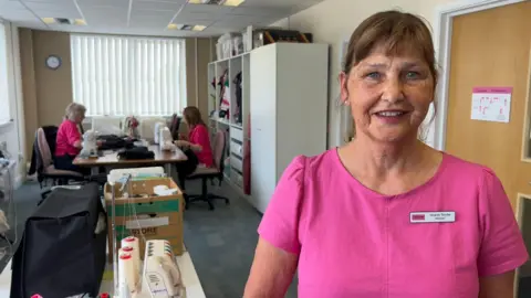 Sharon Tombs is wearing a pink top and a name badge and smiling at the camera. In the background two women are working on sewing machines.