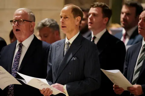 Reuters Men in suits stand singing from printed hymn sheets during. The central man wears a dark pinstripe double-breasted suit with a patterned tie and pocket square. Several other men are visible behind him, also holding service booklets and singing. The setting has warm lighting and stone architecture, giving a ceremonial atmosphere.