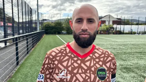 Osama Almahmoud, a man with a shaved head and a thick brown beard, looks at the camera while standing on a football pitch. He is wearing a beige and brown patterned football top with an orange collar and a black EMSONI badge. 