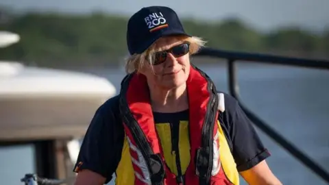 A blonde woman in dark glasses, T-shirt and life jacket wearing a baseball cap which reads 'RNLI 200'. 