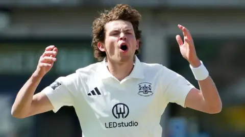 Henry Brookes, wearing a white Adidas cricket shirt with the Gloucestershire county logo, opens his mouth and puts his hands up by the side of his head after bowling a ball in the County Championship Division Two game against Lancashire