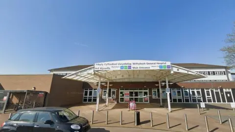 The outside of the front of a hospital. It is a brown brick building with a white canopy at the front and white framed windows. In front of the building are several bollards, and a black car parked on the road.
