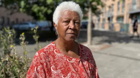 The image shows a middle-aged woman standing in Butetown. She is looking at the camera. She is wearing a red top. It is a sunny day.