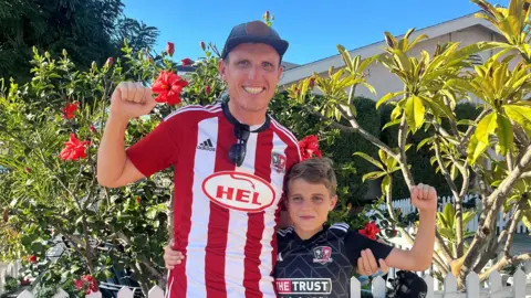 George and Charlie standing together. They are both holding one arm up in the strong position and wearing Exeter City shirts. George is wearing a white and red and top while Charlie is wearing a black and white shirt. There are plants and trees behind them and it is a sunny day.
