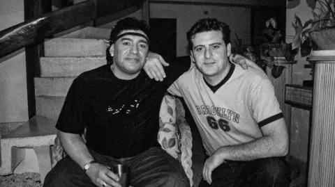 Maradona wearing a black top holding a drink with his arm around Adam Docker who is wearing a light-coloured top. They are inside a building.