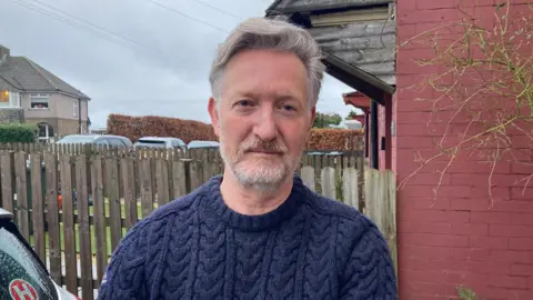 A man wearing a blue cable-knit jumper standing in front of a faded wooden fence and red-brick wall 