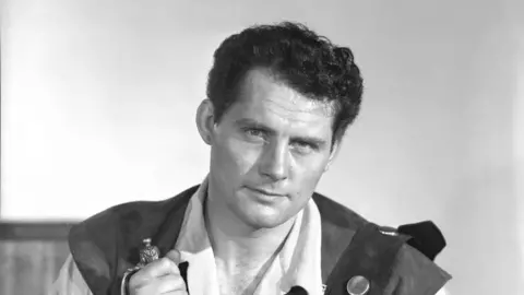 Getty Images Robert Shaw in black and white wearing a white shirt and dark tabard in an 18th century style. He has dark hair and is staring moodily, though looking a little amused, down the camera. In his right hand is a sword.