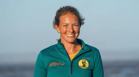 RWREDMANPHOTOGRAPHY A head and shoulders images of a woman with a green top on over a yellow top. She is standing outside, seemingly on a seafront somewhere, smiling and looking into the camera.