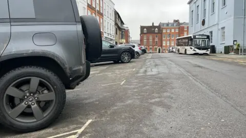 ELLEN KNIGHT/BBC A photograph looking down the road, with the back half of a dark grey vehicle visible on the right. The parking bays are demarcated by white spray-painted lines. The buildings are Georgian, three-storey homes, with a large theatre on the right of the street, and a white single-decker bus idling outside it. The sky is grey and overcast. 