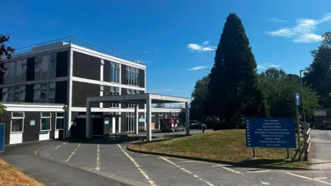 An external view of the maternity unit at North Devon District Hospital. It is a brown building with white features. Either side is grass areas, and on the right a blue sign with directions to different departments.