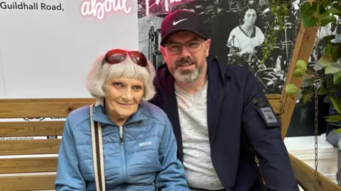 A woman in a blue puffer jacket and jeans sits on a wooden bench. Next to her is a man in a navy coat, a white jumper and grey jeans. They are looking at the camera and on the left hand side are lots of flowers. 