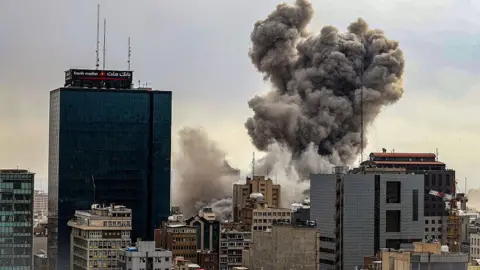 Getty Images A large plume of dark grey and brown smoke billows upward between high-rise buildings in a dense urban area. A glass-fronted tower block is visible to the left. Construction cranes and telecommunications masts can be seen among the rooftops.
