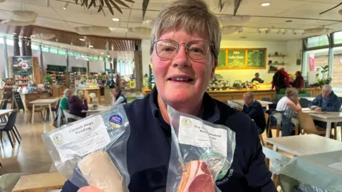 Sally Lugg holding a packet of bacon and a packet of meat. She has glasses and dark grey hair. She is in a cafe setting.